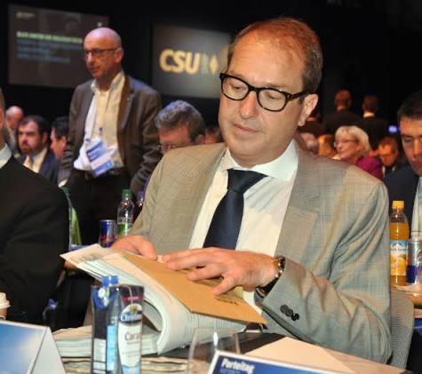 Alexander Dobrindt in a light grey jacket reading documents at a CSU party conference, with delegates in the background