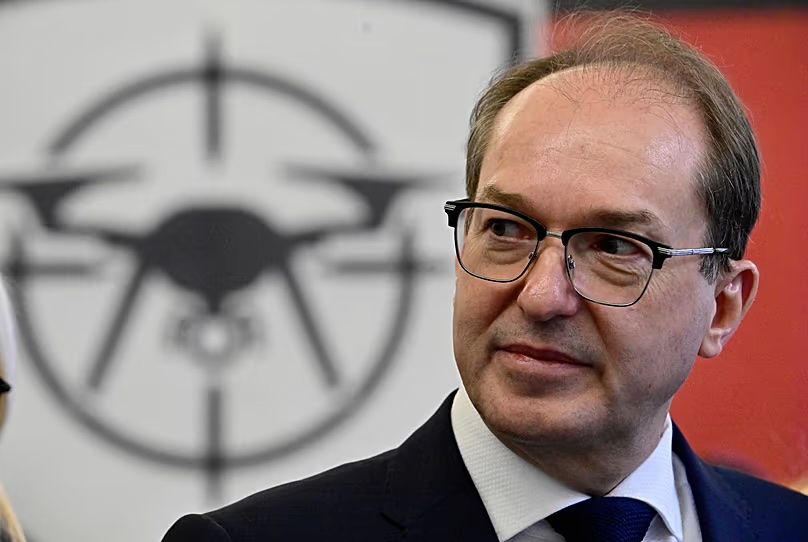 Close-up of Alexander Dobrindt in a dark suit, with a drone-defence emblem showing a drone in crosshairs behind him