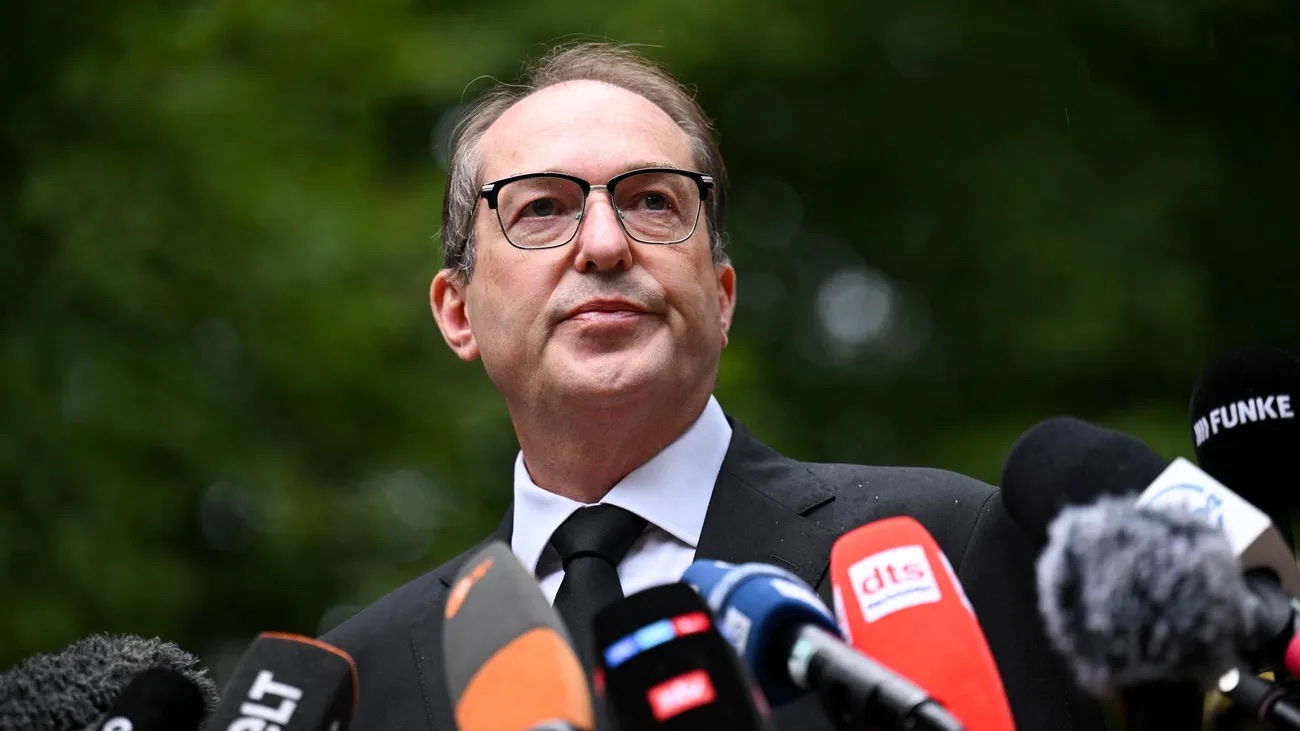Alexander Dobrindt in a dark suit and black tie speaking outdoors to a bank of press microphones
